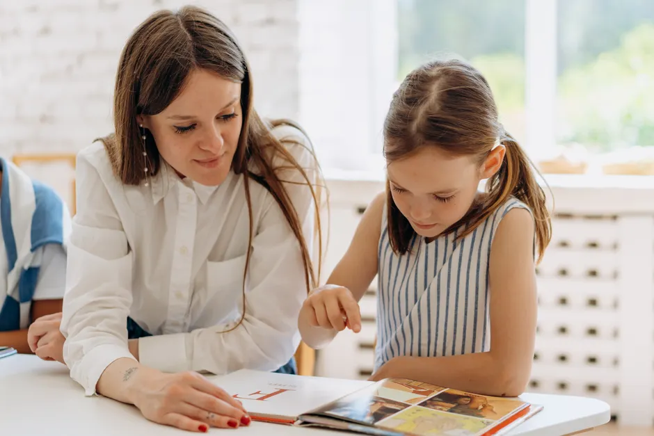 A teacher guides a young girl reading a book indoors, emphasizing learning and connection.