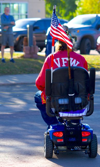 A veteran on a mobility scooter carrying a US flag outdoors, wearing a VFW jacket.