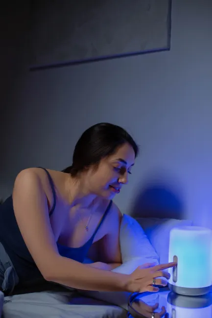A woman in a bedroom setting interacts with a calming blue light lamp, creating a peaceful nighttime atmosphere.