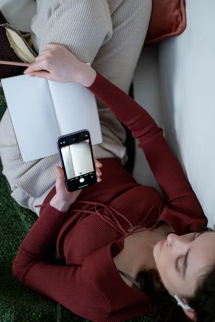 A woman lying down holding a smartphone and an open notebook, viewed from above.