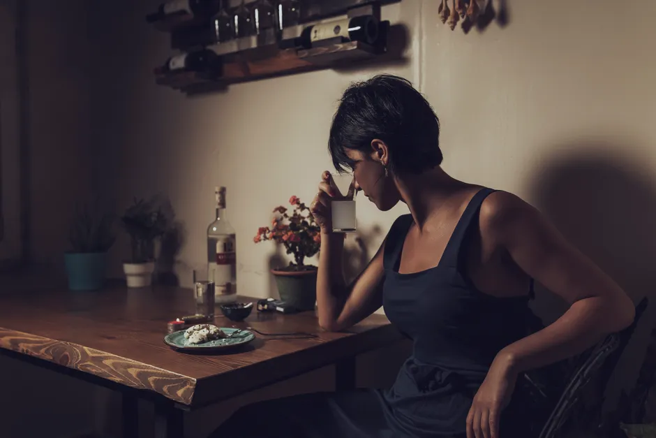 A woman seated alone at a table holding a drink in a dimly lit room, evoking solitude.