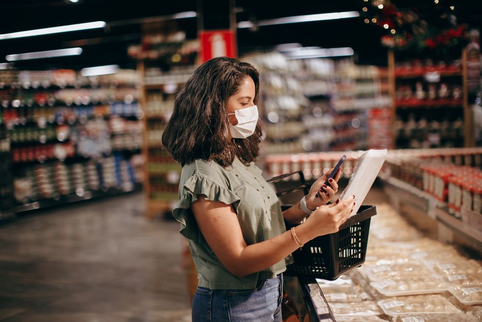 Abus économique dans le couple : quand l’argent devient un outil de contrôle A woman shopping in a supermarket, wearing a face mask and using her phone to check products.