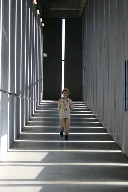 A young boy walking down a sunlit modern hallway, casting dramatic shadows.