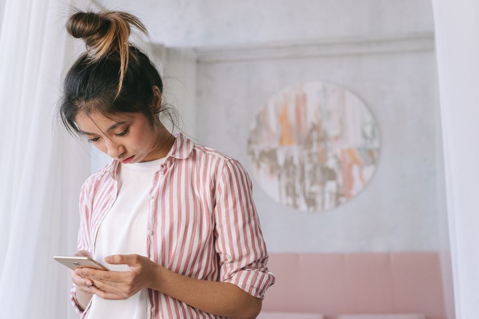 A young woman in a striped shirt using her smartphone indoors with soft decor and a relaxed atmosphere.