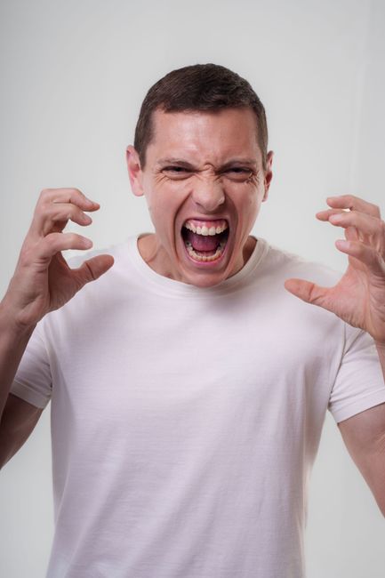 An adult man in a white t-shirt expressing strong emotions by shouting indoors.