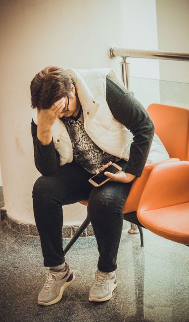 An anxious woman sits in a waiting room, holding her smartphone and resting her head on her hand.