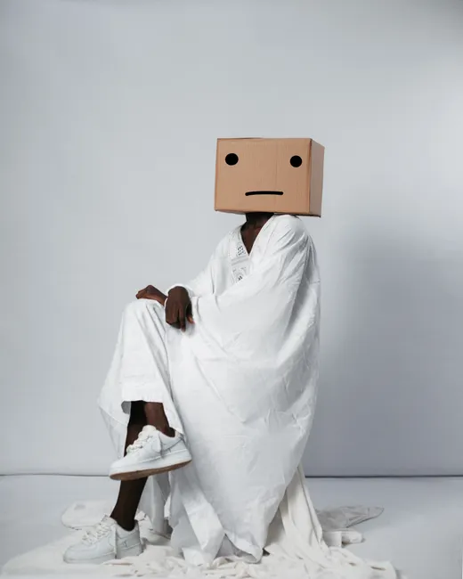 Artistic photo of an anonymous individual wearing a cardboard box mask and white robe, seated.