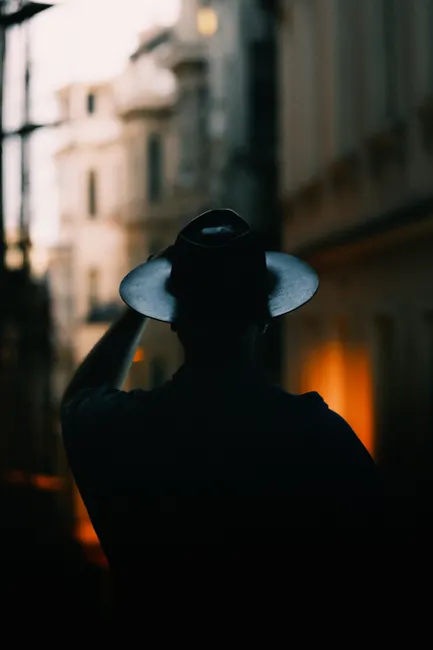 Backlit silhouette of a person wearing a fedora hat, capturing a mysterious urban evening scene.