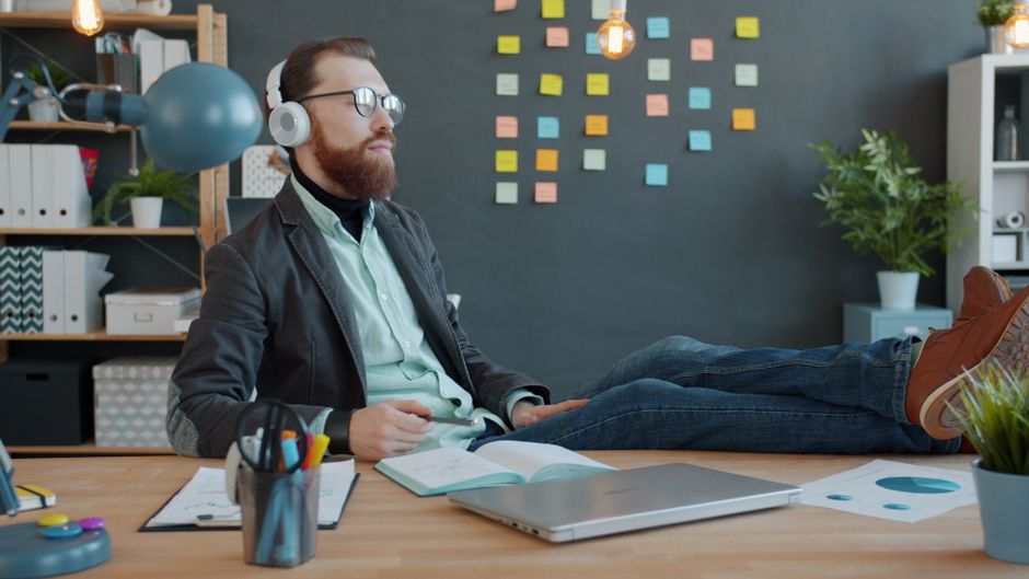 Mindfulness au travail : ce que la pleine conscience change, et ce qu’elle ne répare pas Casual office worker wearing headphones, relaxing with feet up on desk. Modern creative workspace with sticky notes and plants.