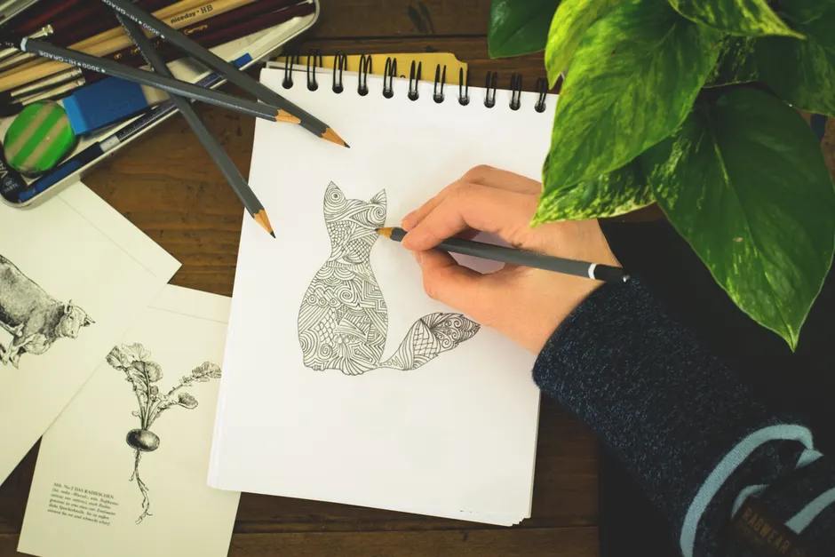 Close-up of a hand drawing a detailed cat design in a notepad surrounded by art supplies and plants.