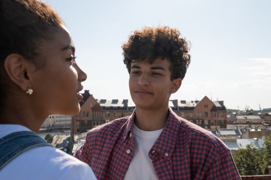Close-up of two teenagers talking on a rooftop with an urban backdrop on a sunny day.