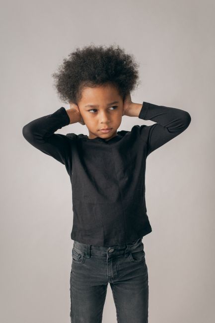 Desperate African American girl in black outfit and Afro hairstyle looking away while covering ears on white background in room