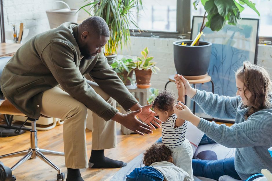 Diverse parents supporting infant learning walking near brother while spending time together at home