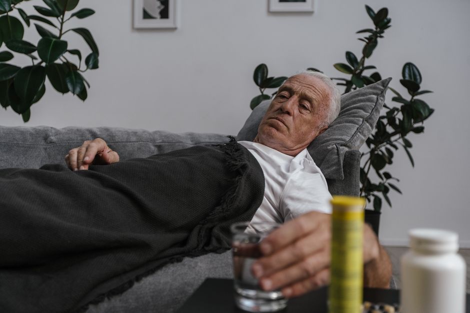 Elderly man lying on sofa with medications, feeling unwell and resting indoors.