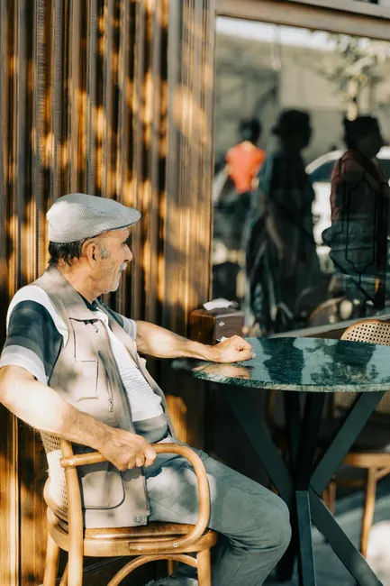 Elderly man sitting alone at a cafe table outdoors, reflecting in the sunlight.