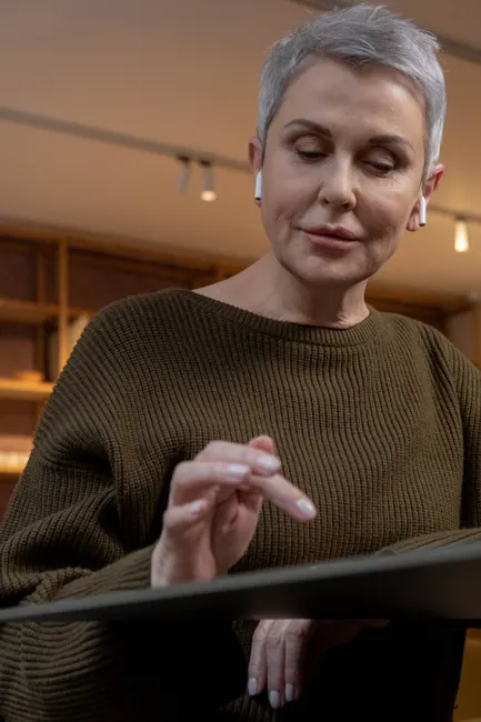 Elderly woman in brown sweater using a tablet with wireless earbuds indoors.