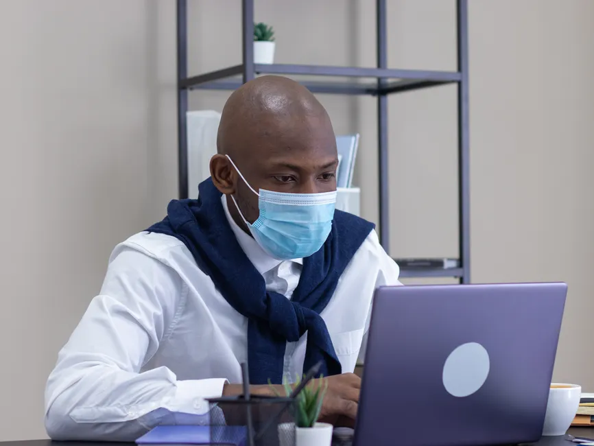 Télétravail ou bureau : ce que la psychologie dit de la productivité Focused office worker wearing a face mask while working on a laptop in a modern workspace.