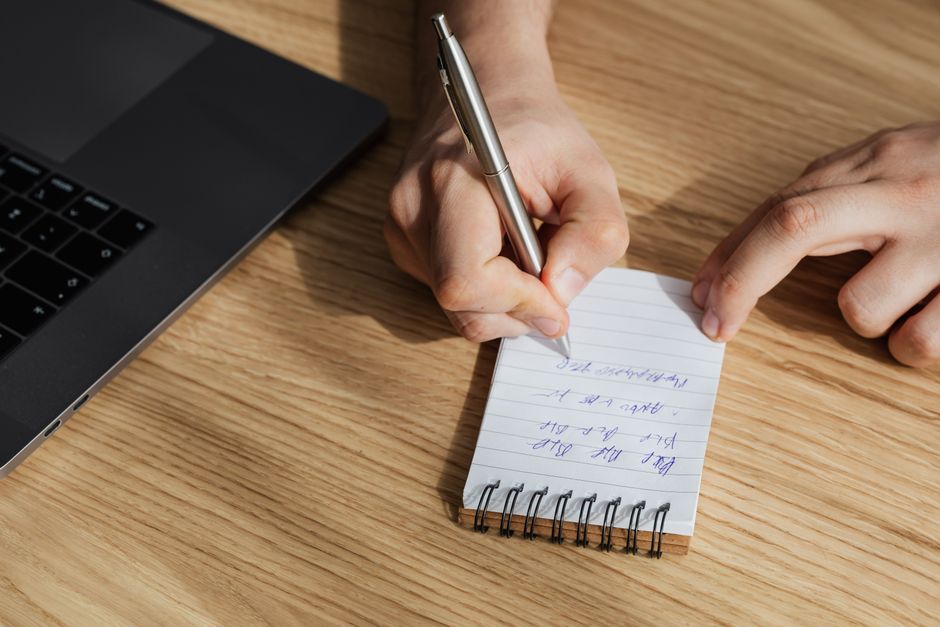 From above of crop unrecognizable man writing out information in spiral notebook while working on business plan on laptop
