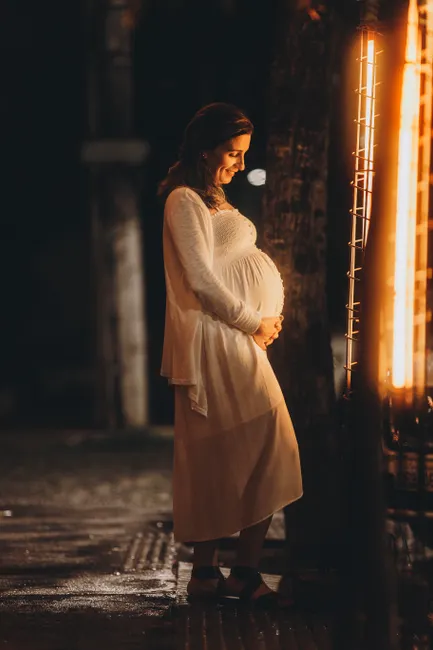 Happy pregnant woman admiring lights on a tree at night, embracing motherhood.