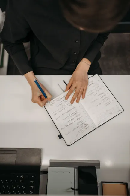High-angle view of a person writing notes in a diary on a white desk, featuring a laptop and stationery.