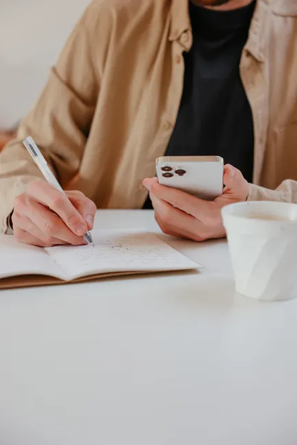 Man taking notes on a notebook while using a smartphone at a desk with a cup of coffee.