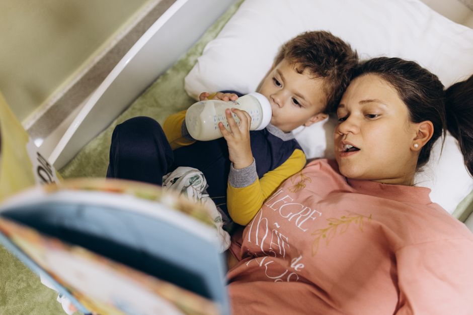 Mother and child lying in bed, bonding over a bedtime story and bottle feeding.