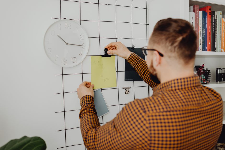 Person arranging notes on an office wall grid beside a clock, creating an organized workspace.