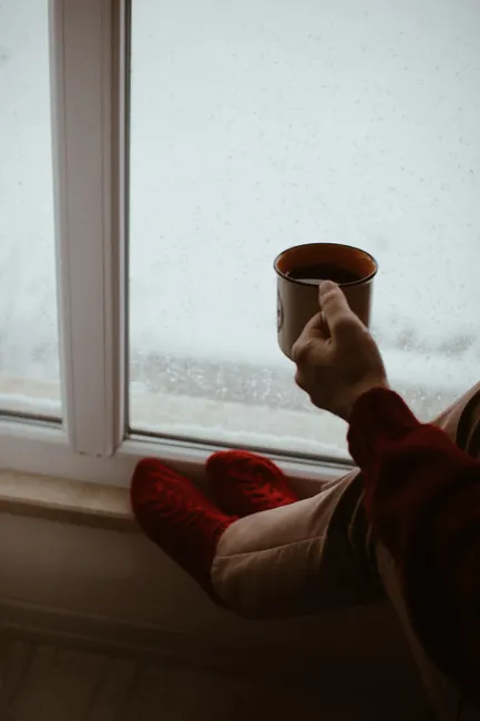 Person holding a coffee mug sitting by a window on a winter morning. Red socks add warmth.