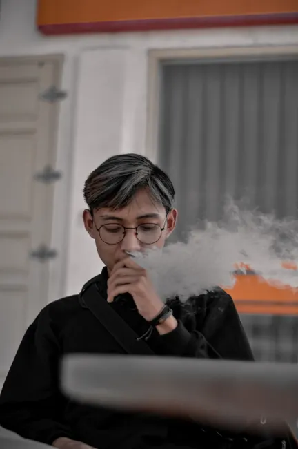 Portrait of a young adult wearing glasses while vaping indoors, producing a visible cloud of vapor.