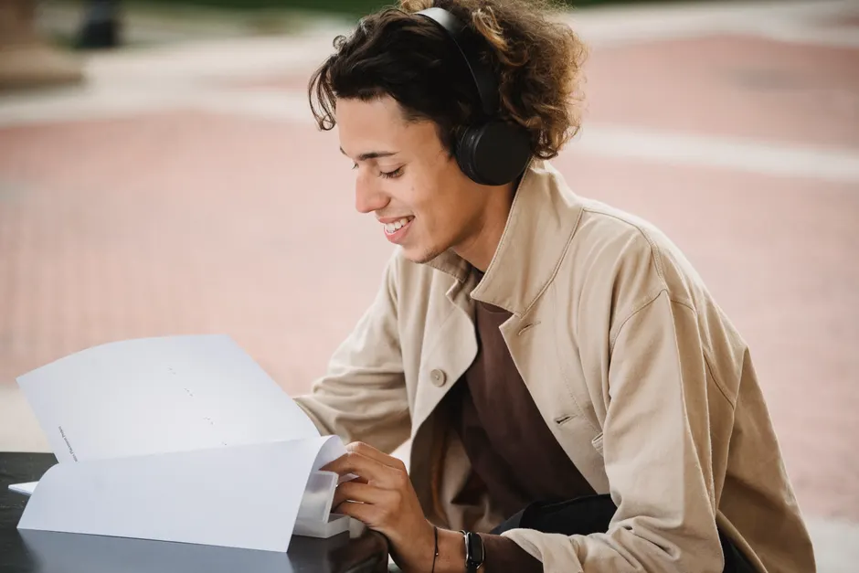 Side view of smiling male student looking through papers with homework task while listening to music in wireless headphones