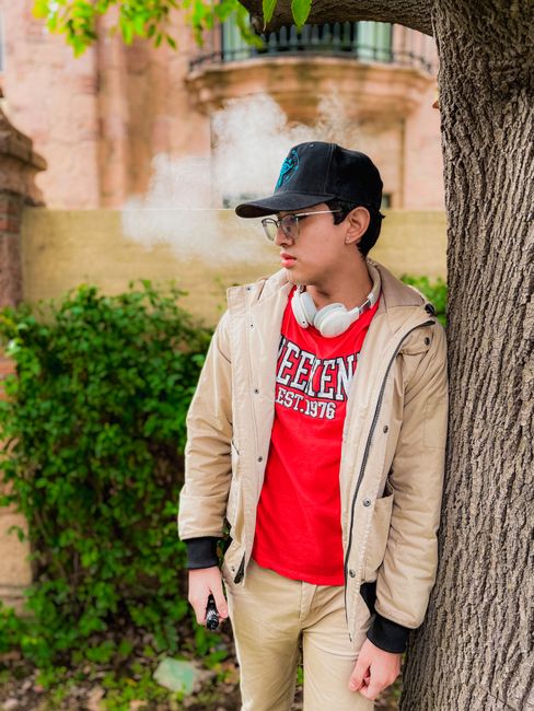 Teenager in a baseball cap and headphones by a tree, looking pensive.