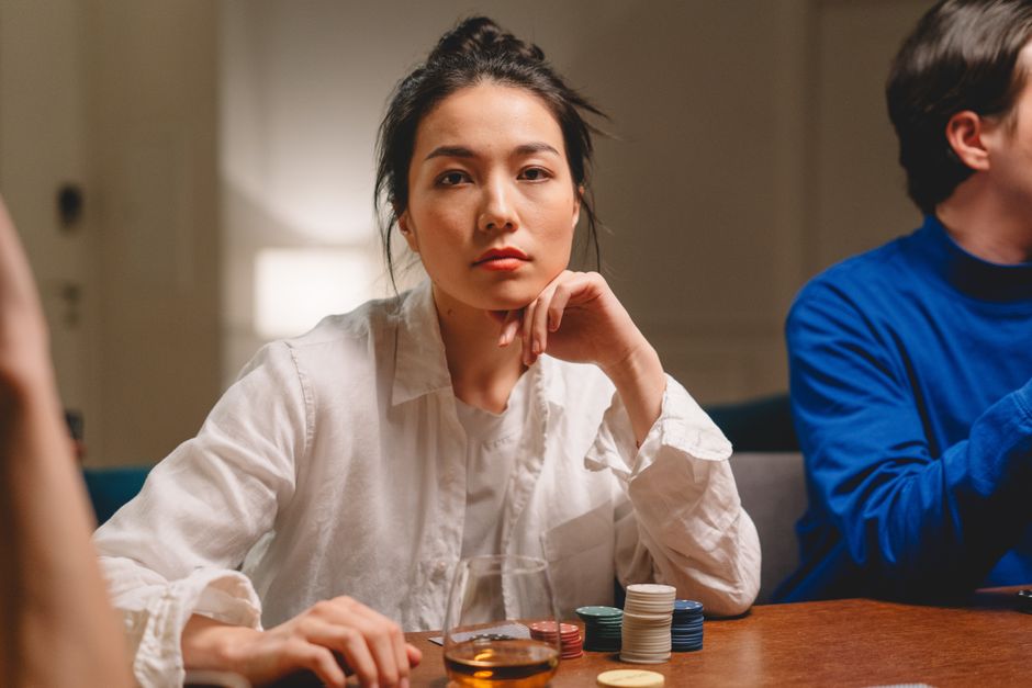 Thoughtful young Asian female in casual clothes sitting at table with gaming chips and whiskey and playing with crop anonymous friends