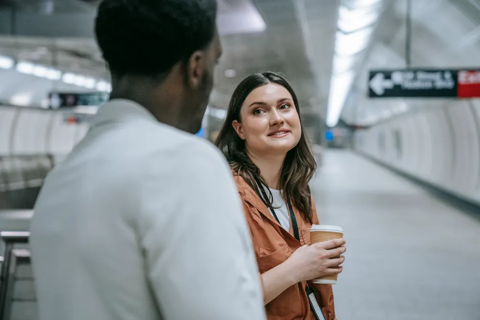 Two adults talking at a subway station platform, holding a coffee cup, during the day.