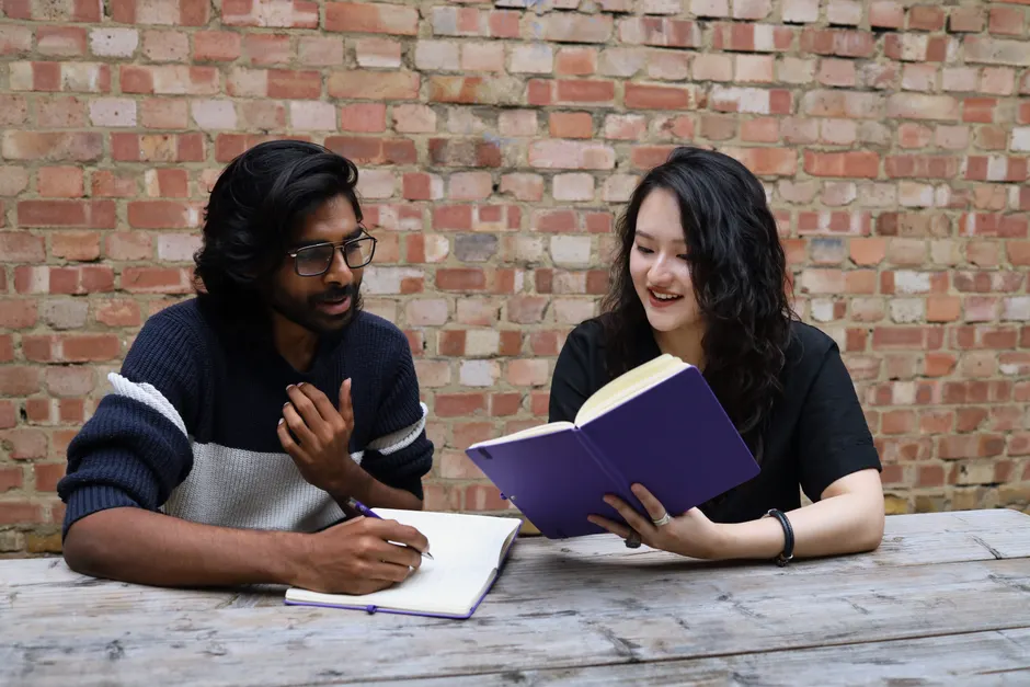 Two young adults enjoying a study session outdoors, discussing and writing in notebooks.