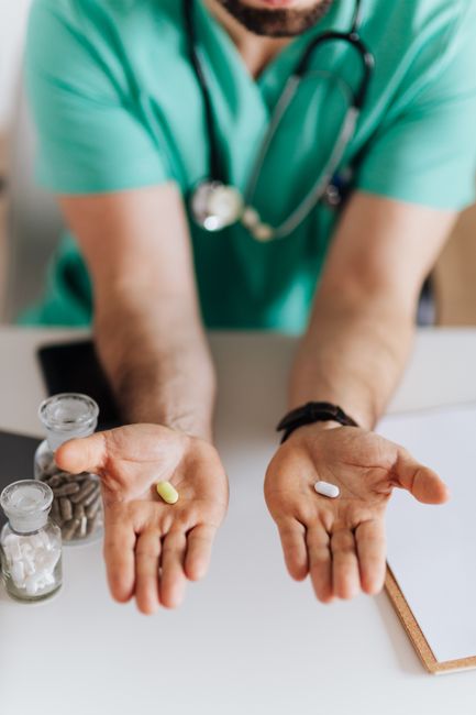 Unrecognizable male doctor wearing medical uniform showing pills to camera while working in modern clinic sitting at table with clipboard and stethoscope