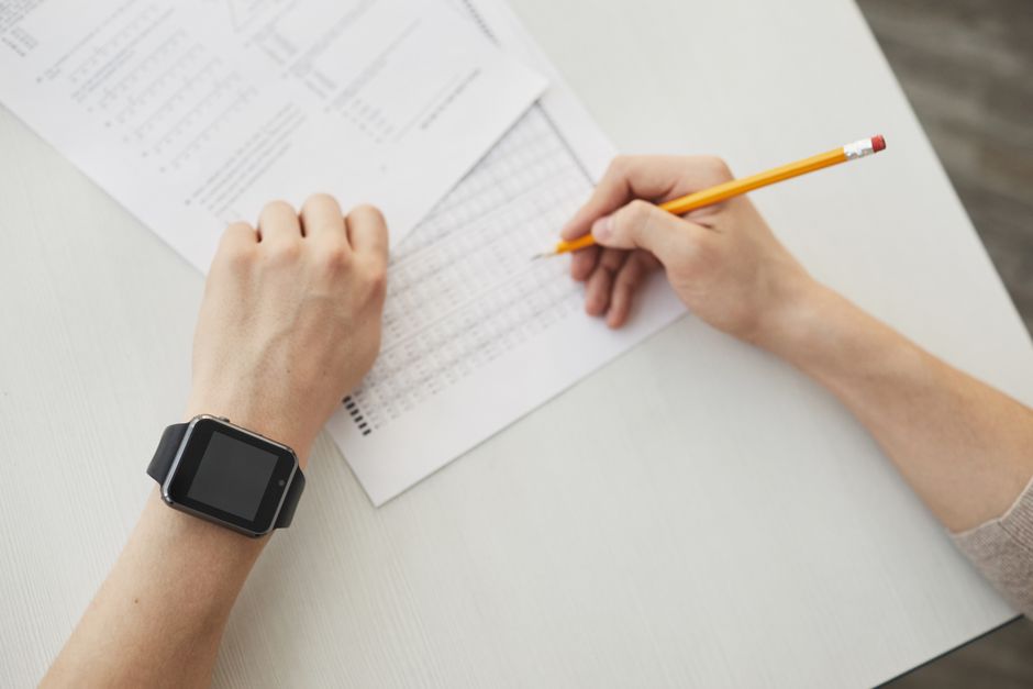 Unrecognizable student writing an exam using a pencil, watch visible on wrist.