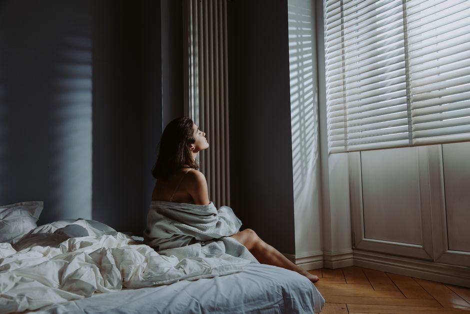 Woman sitting on bed in cozy bedroom, wrapped in a blanket with sunlight streaming through blinds.
