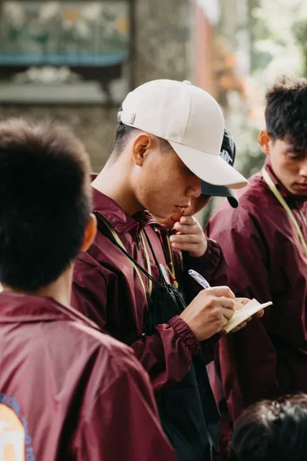 Young adults in maroon jackets gather outdoors, engaging in conversation and note-taking.