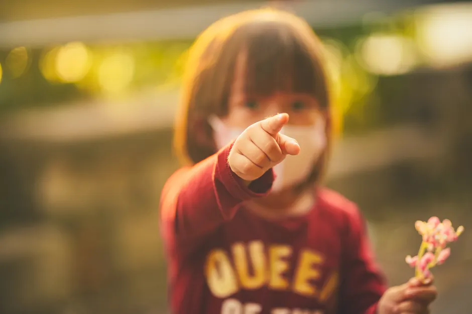 Young child wearing a mask, playfully pointing forward in a warm outdoor setting.