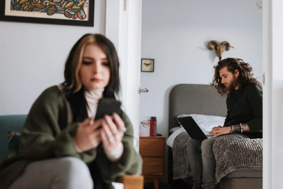 Young hipster male surfing internet on netbook on bed against female partner with cellphone in house